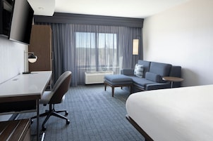 Habitación de hotel con cama, escritorio, silla, sofá, mesita, televisión y ventana con cortinas.