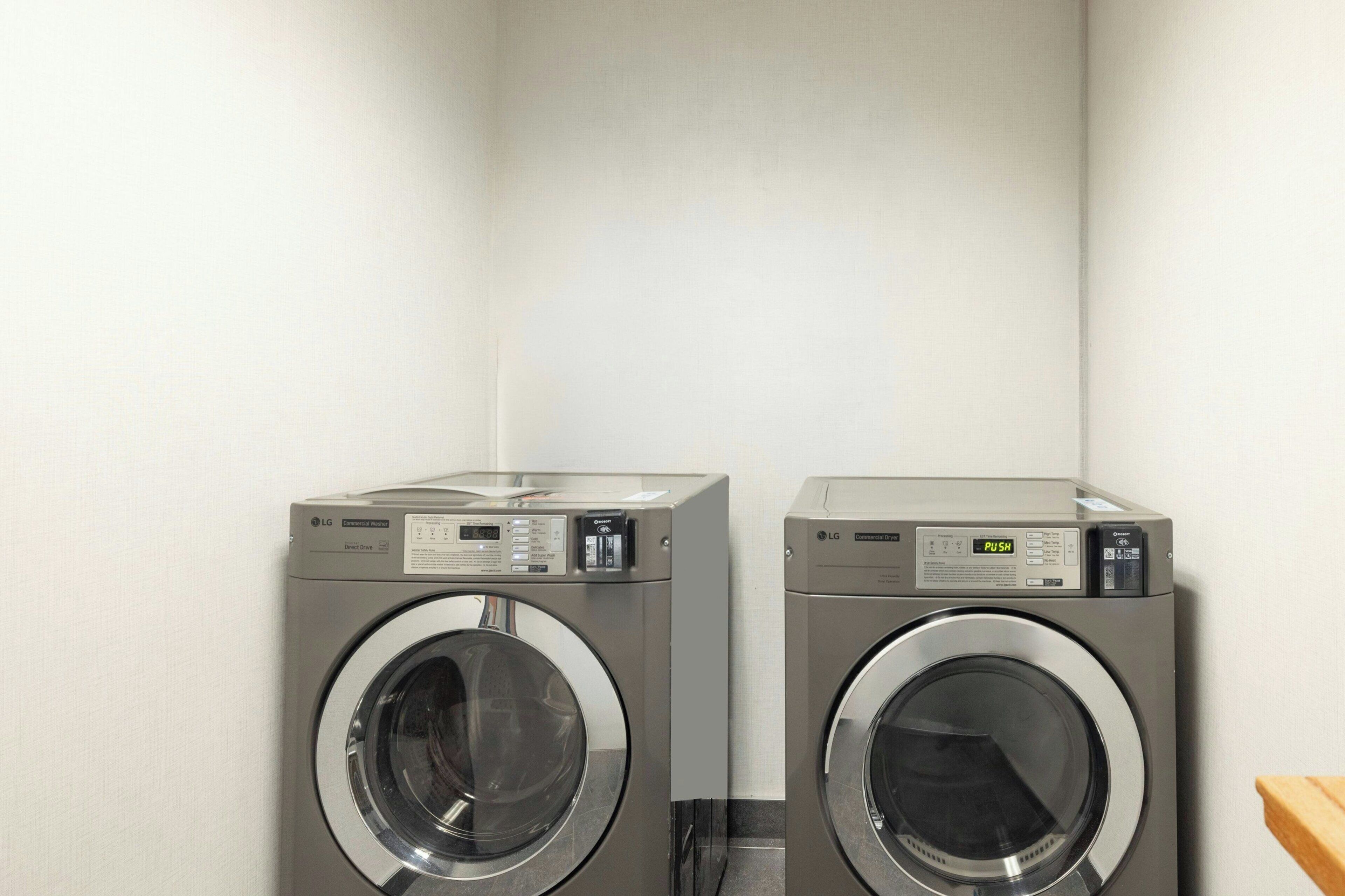 Two commercial washers in a corner with a plain wall and a wooden bench.