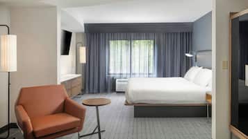 Une chambre d’hôtel avec un lit, une chaise, une petite table, un bureau et une fenêtre avec des rideaux.