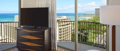 A hotel room with a large TV, a small round table, and a sofa. The room has a balcony with a view of the ocean and buildings.