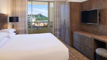 A hotel room with a large bed, a flat-screen TV mounted on the wall, a wooden dresser, and a view of a mountainous landscape through the sliding glass door.