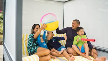 Une famille est assise sur un lit, recouvert d’une couverture à rayures, avec un ballon de plage et un jouet rouge.