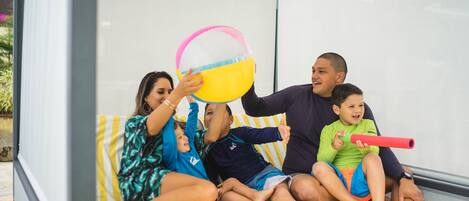 A family sitting on a bed with a striped blanket, a beach ball, and a red toy.