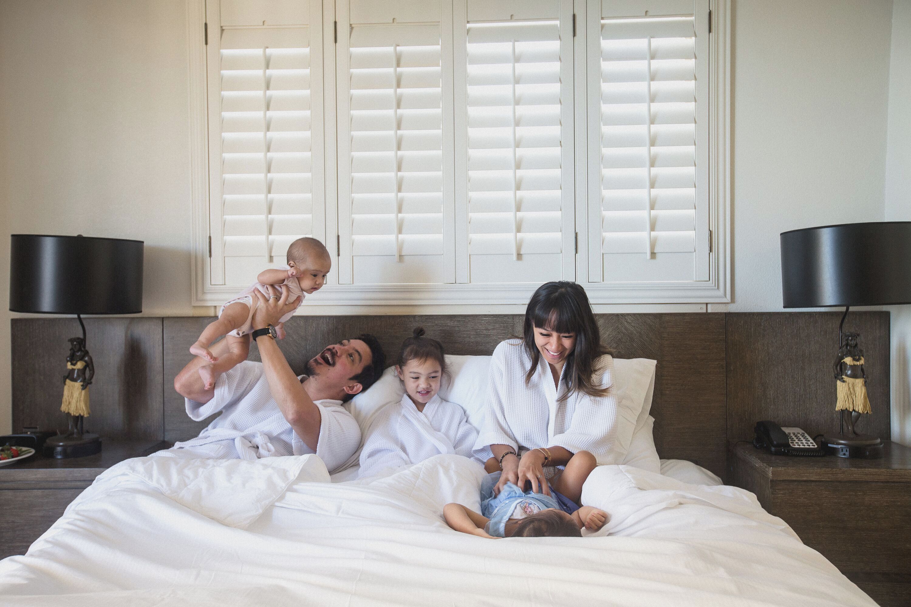 A family of four in a hotel room with a bed, nightstands, and a window with shutters.