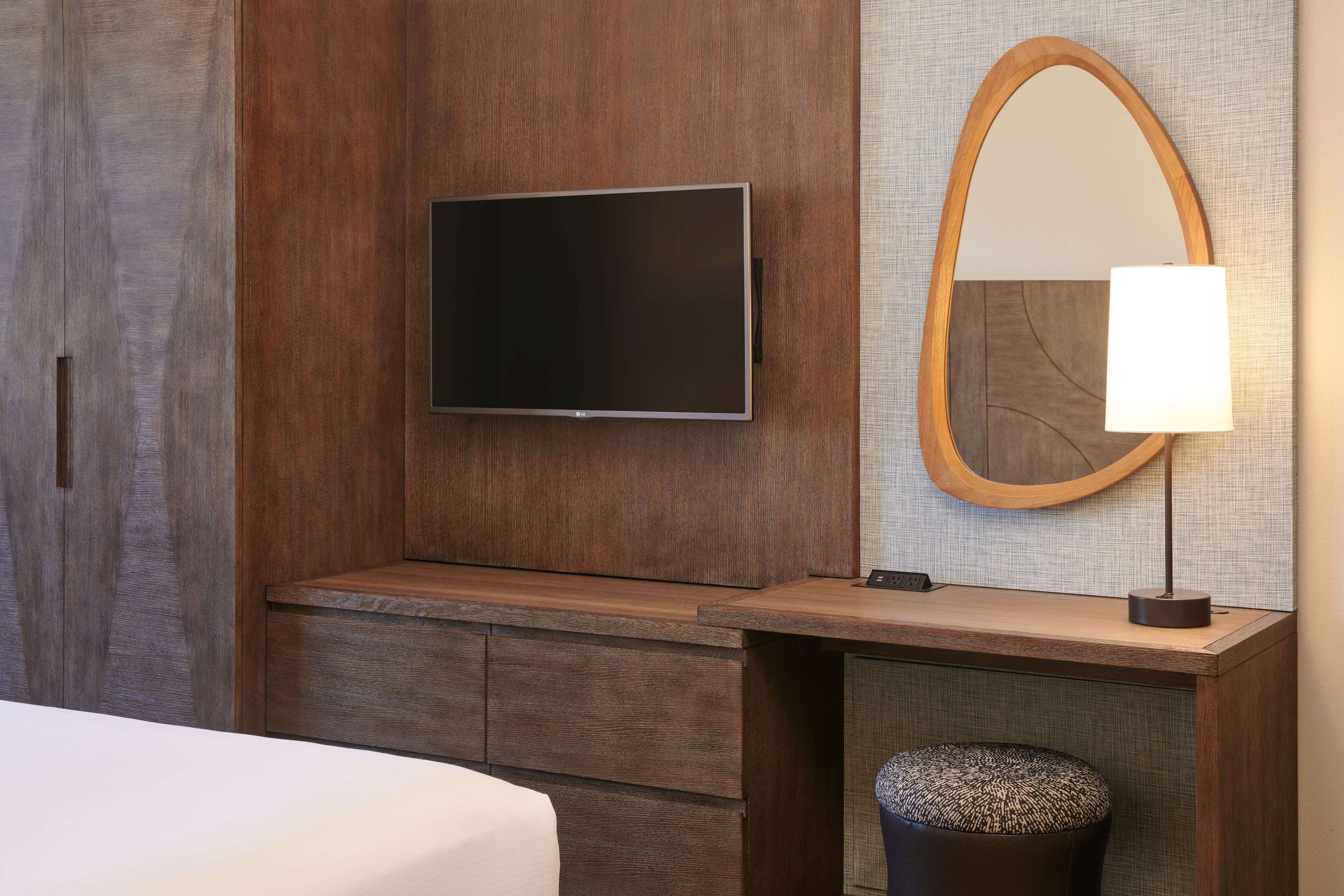 A hotel room with a flat-screen TV mounted on a wooden wall, a wooden bench, a mirror with a wooden frame, a lamp, and a small round ottoman.