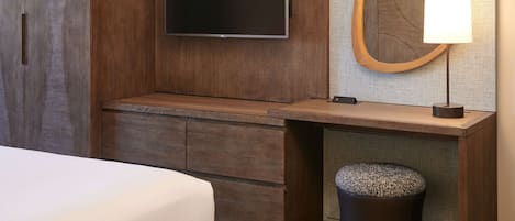 A hotel room with a flat-screen TV mounted on a wooden wall, a wooden bench, a mirror with a wooden frame, a lamp, and a small round ottoman.