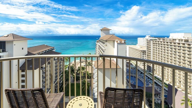 A balcony with wicker chairs, a clock, and a view of the ocean and hotel buildings.