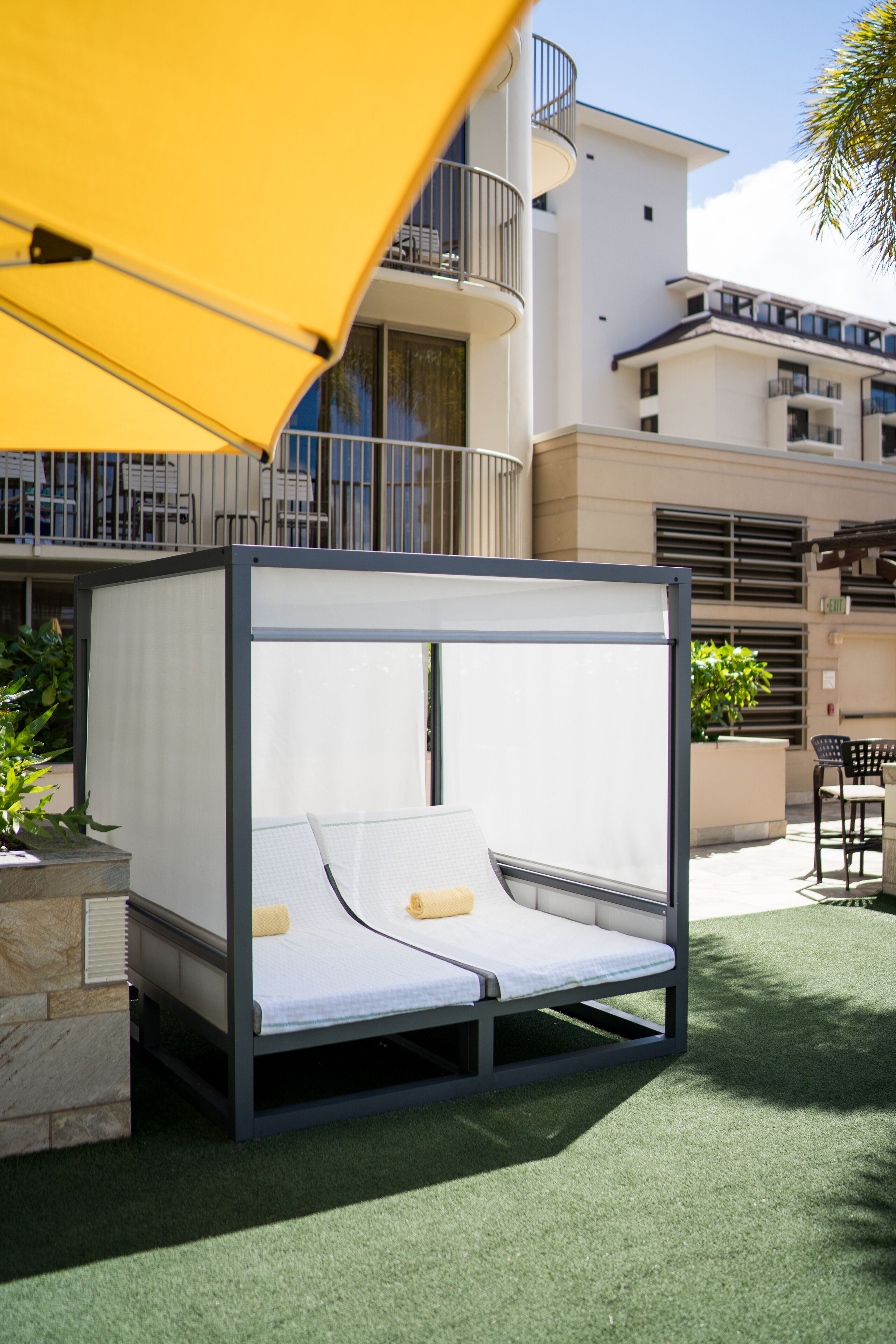 A modern outdoor bed with a canopy, set on artificial grass, with a building in the background.