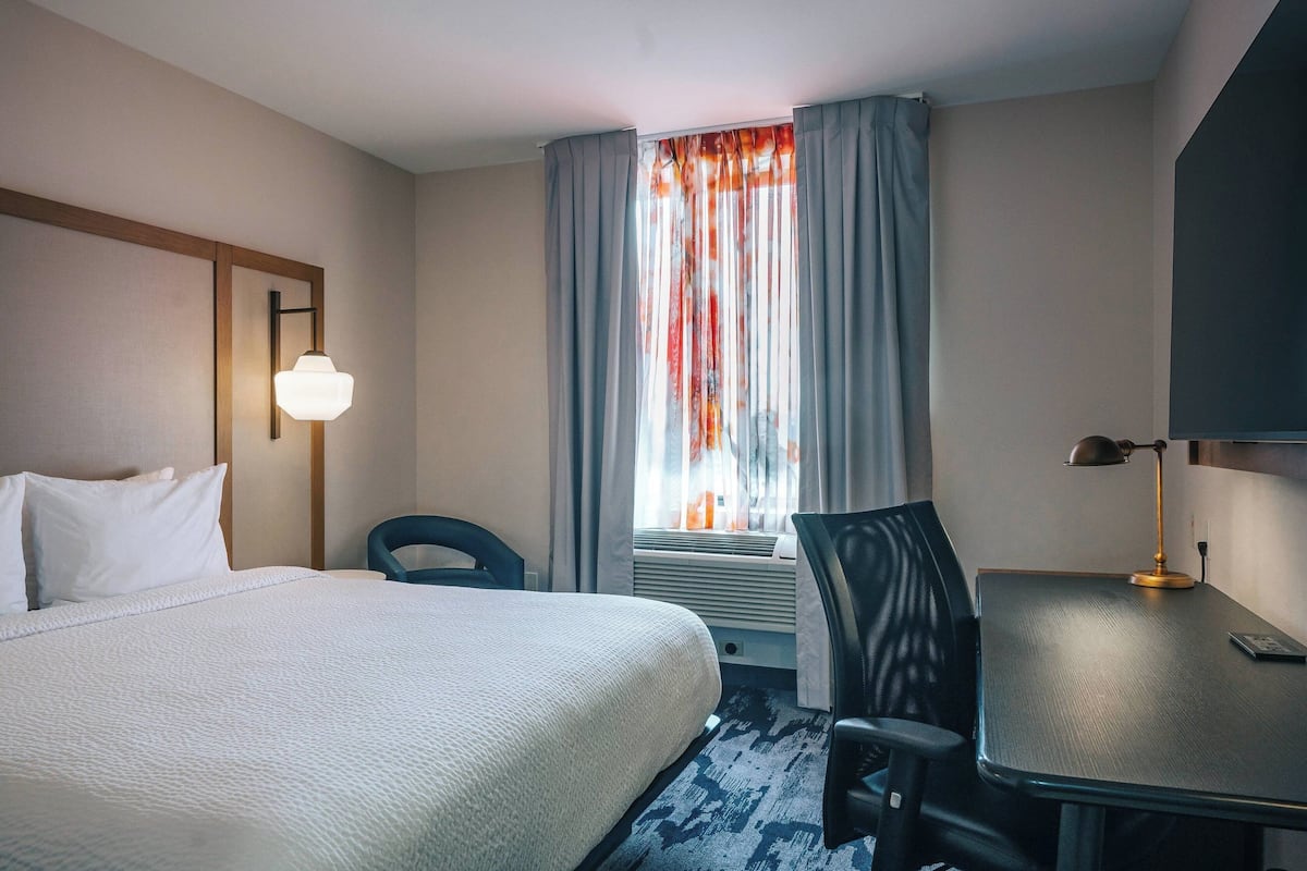 A hotel room with a large bed, a desk, a chair, a lamp, and a window with curtains.