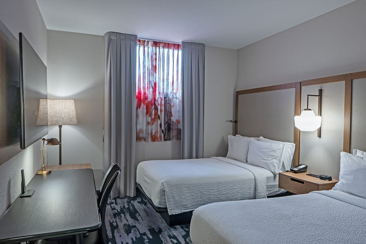 A hotel room with two beds, a desk, a lamp, a nightstand, and a window with curtains.