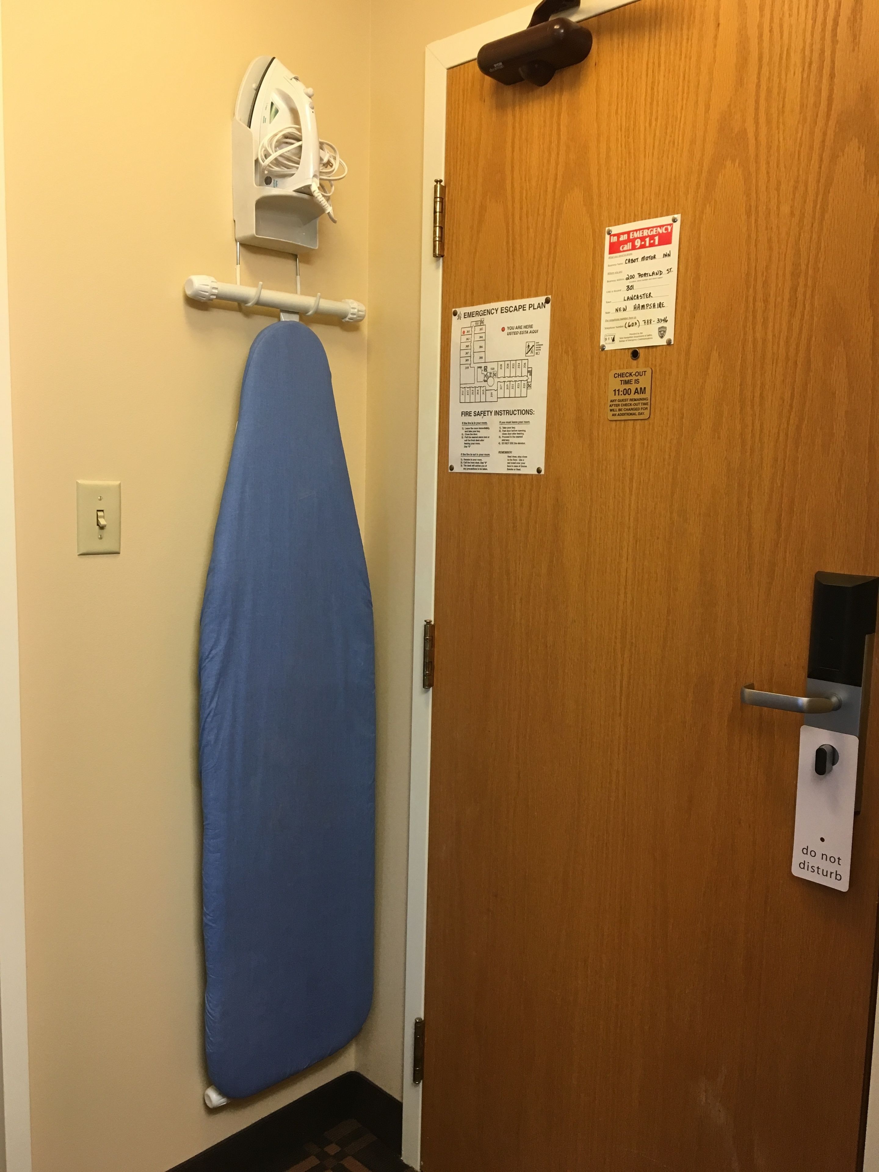 A room with a wooden door, a blue ironing board, a white iron, and emergency exit instructions.