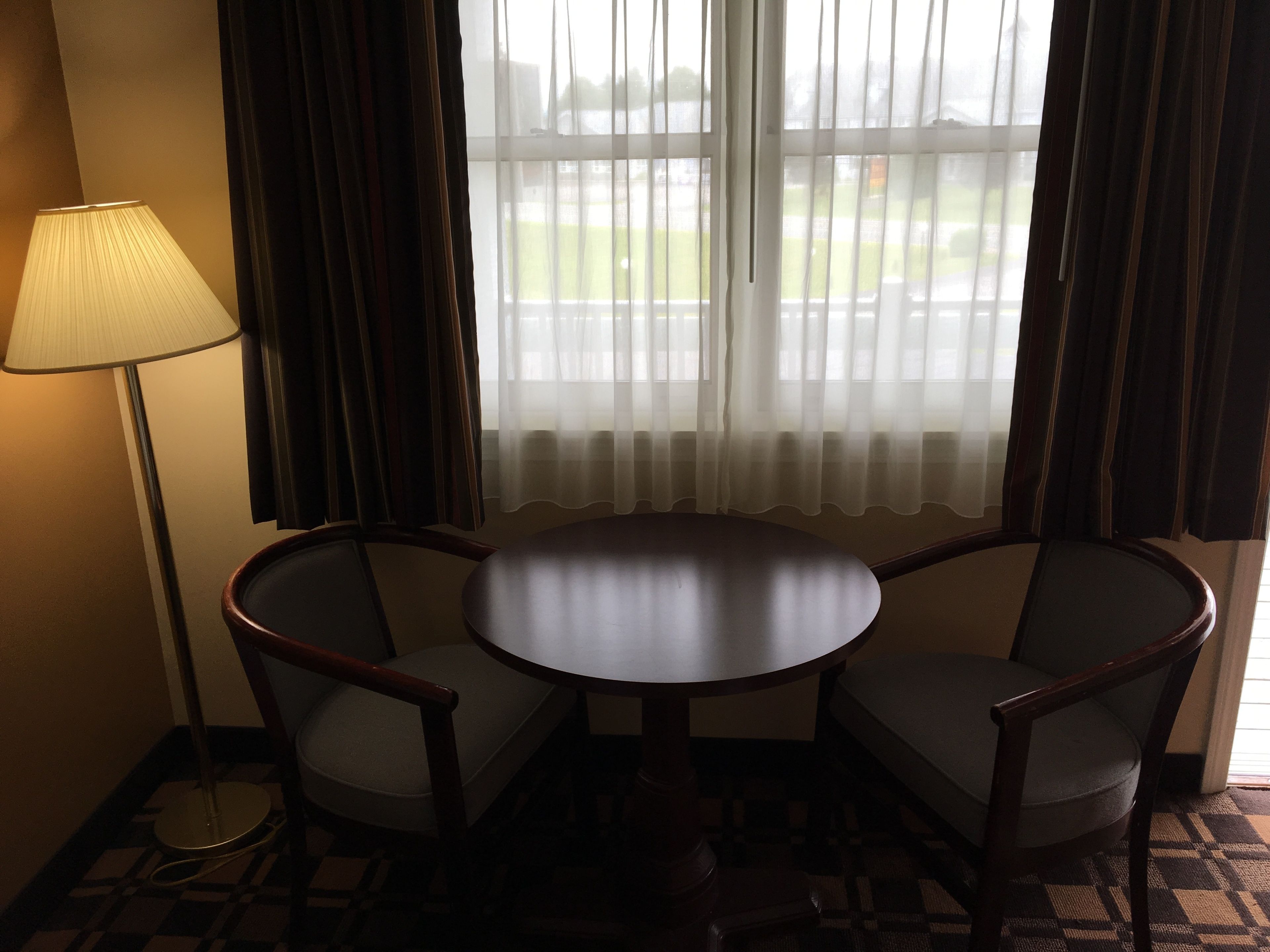 Ein Hotelzimmer mit einem runden Tisch, zwei Stühlen, einer Stehlampe und einem fenster mit Vorhängen.