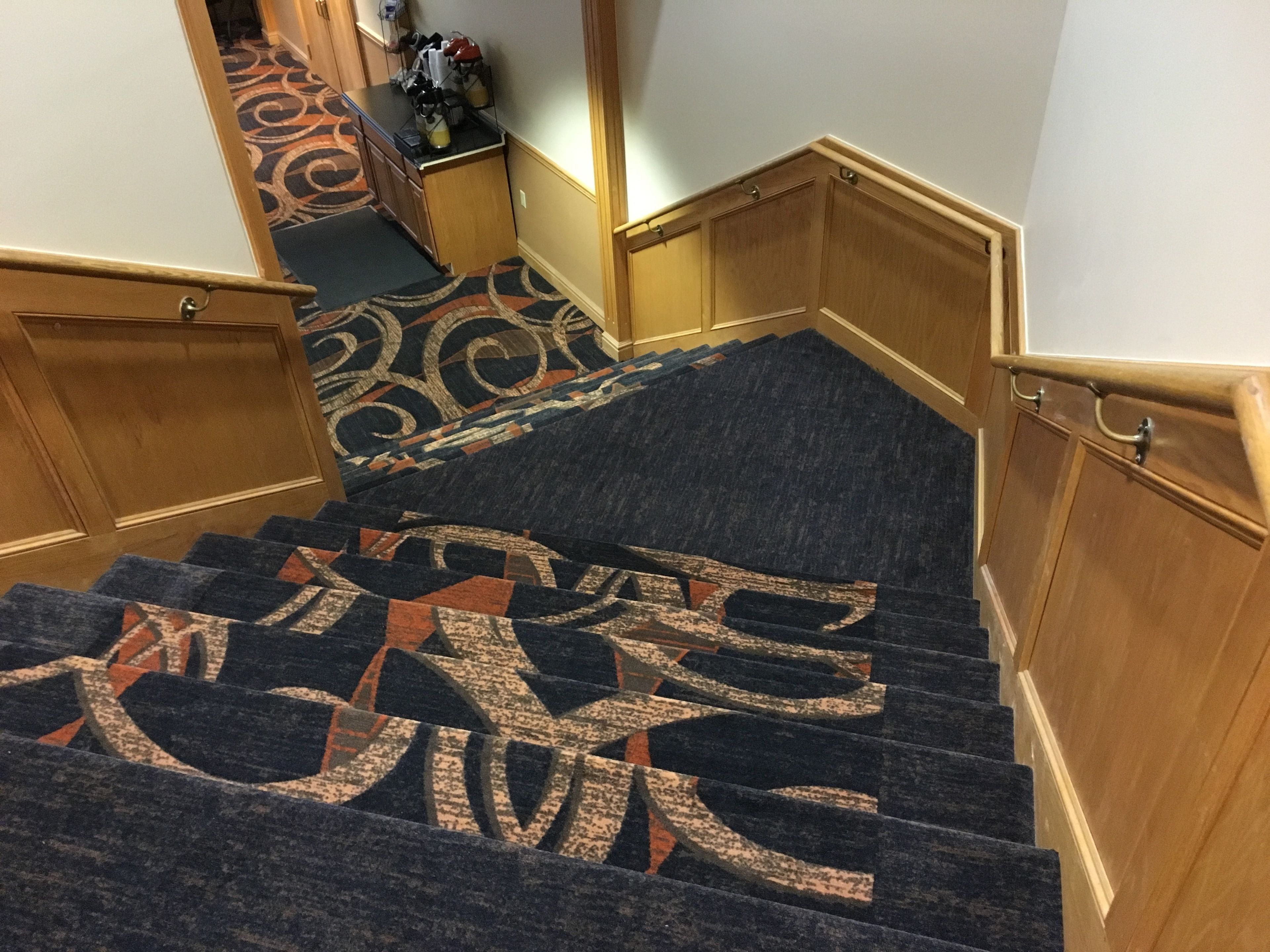 Un escalier recouvert de moquette, orné d’un motif géométrique, avec des rampes en bois et un palier également recouvert de moquette, agrémenté d’un tapis à motifs.
