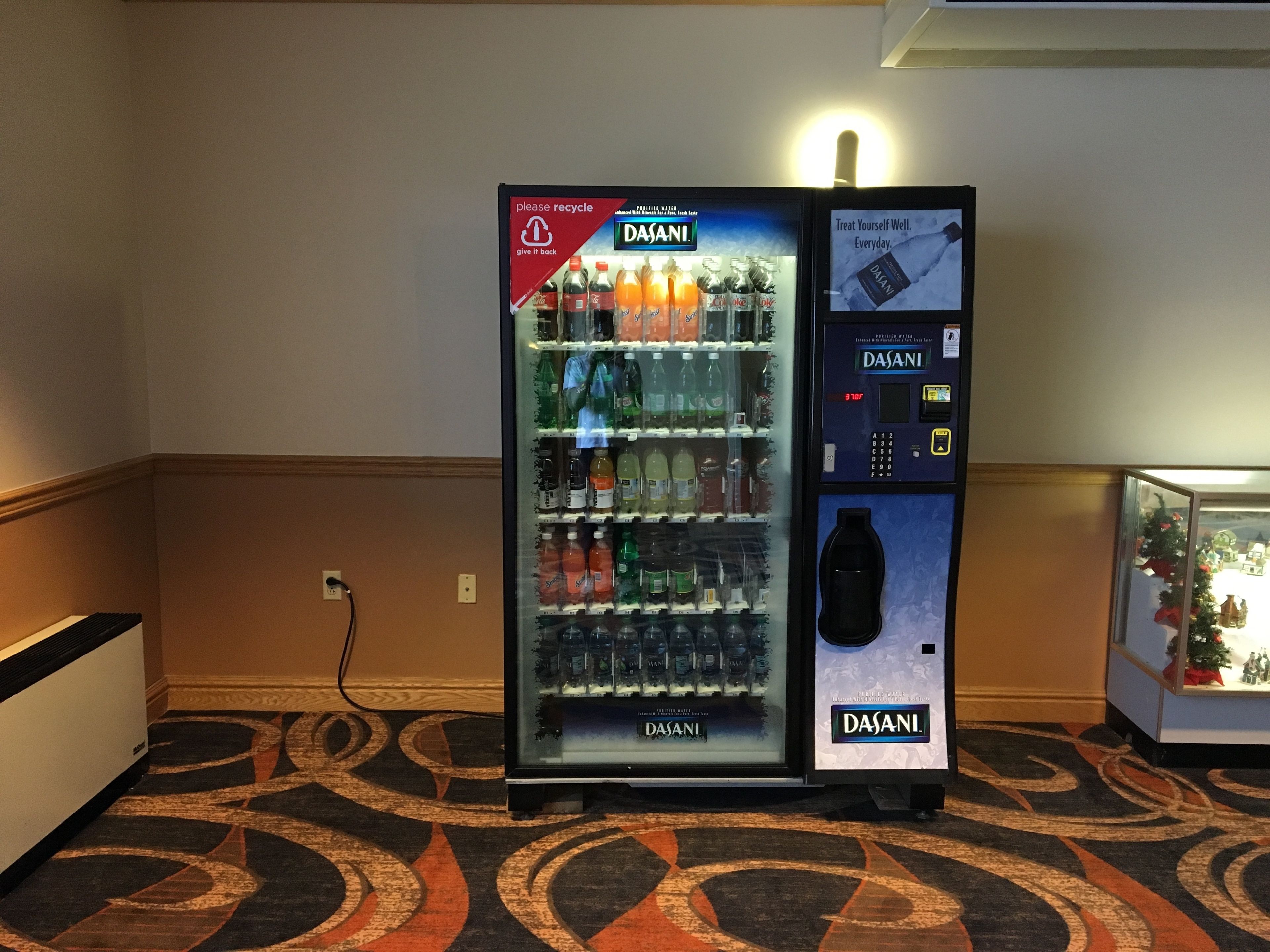 Un distributeur d’eau Dasani doté d’un terminal de paiement numérique, situé dans le hall d’un hôtel, avec un tapis à motifs et une petite décoration de Noël.