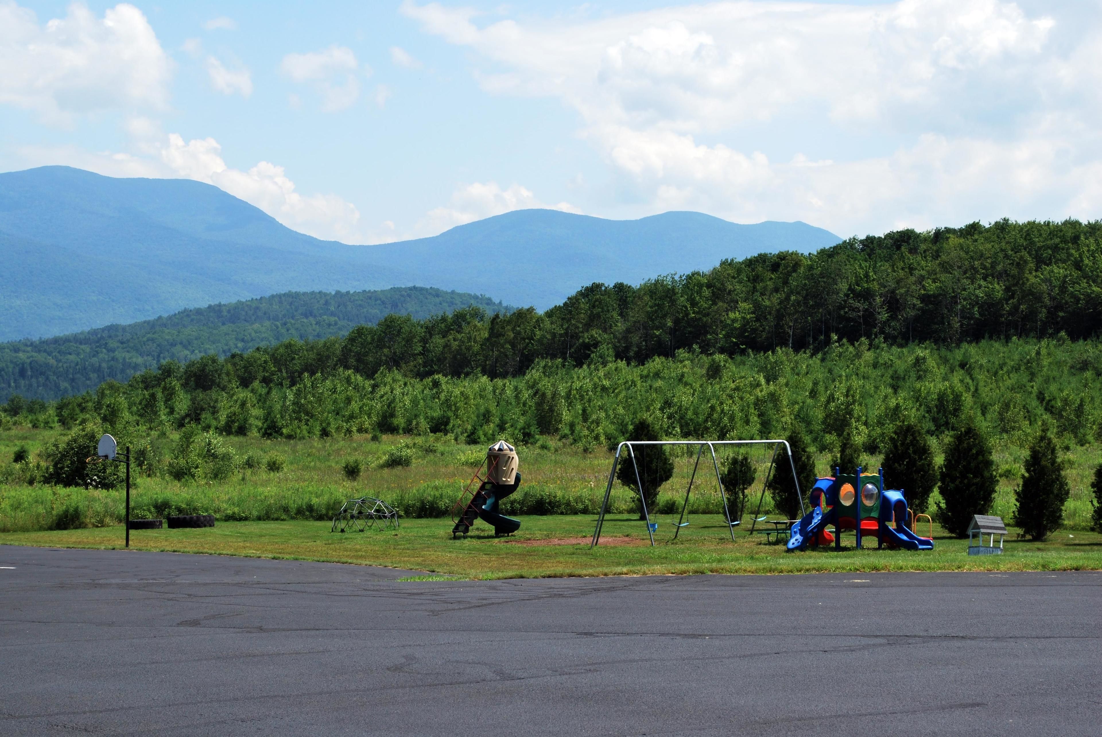 A playground with a slide and swings, set against a backdrop of green hills and mountains.