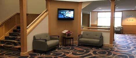 A lobby with a carpeted floor, two armchairs, a small table, a television on the wall, and a staircase.