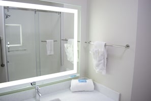 Un baño con un espejo grande, cabina de ducha de vidrio, toallero y lavamanos con encimera blanca.