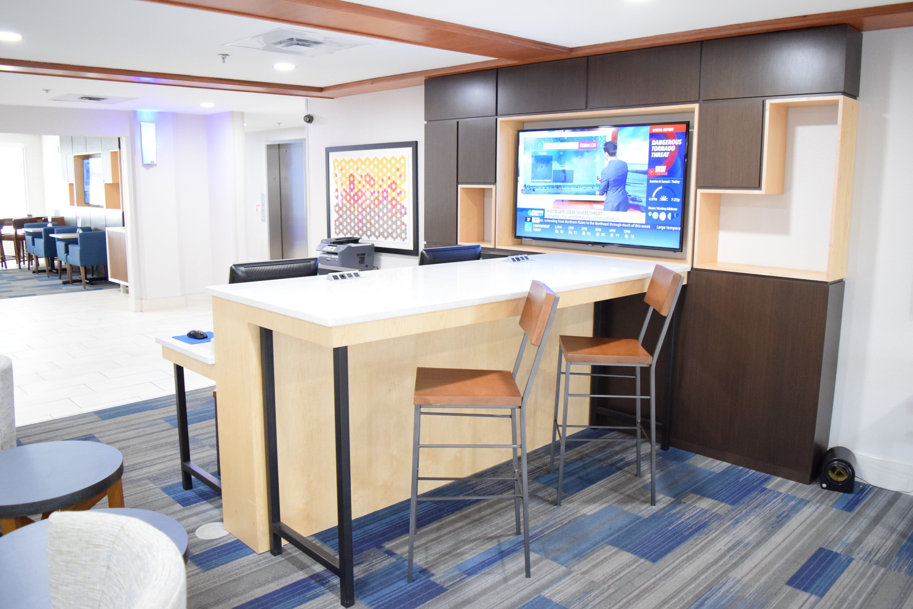 A modern hotel lobby with a reception desk, seating area, and a television displaying a weather forecast.