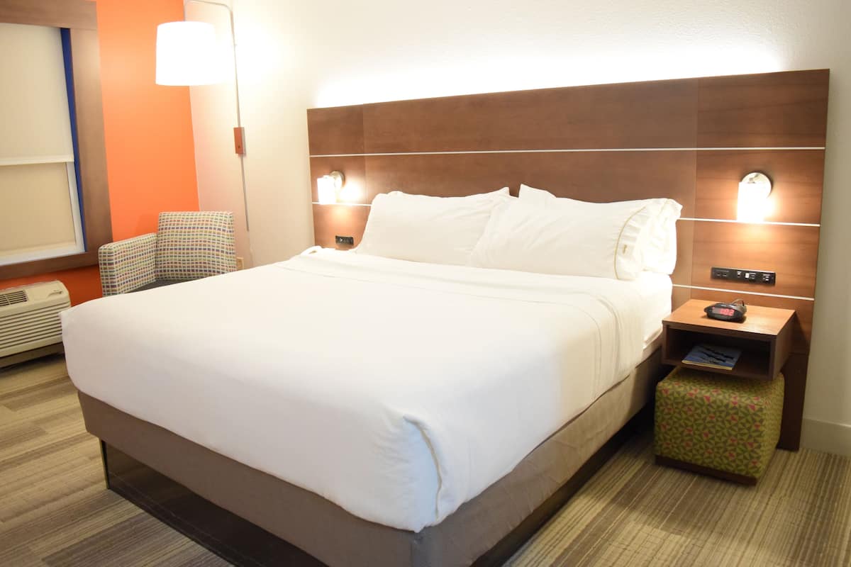 Una habitación de hotel con una cama grande, dos lámparas de noche, una mesita con un reloj, una silla y un aire acondicionado instalado en la pared.