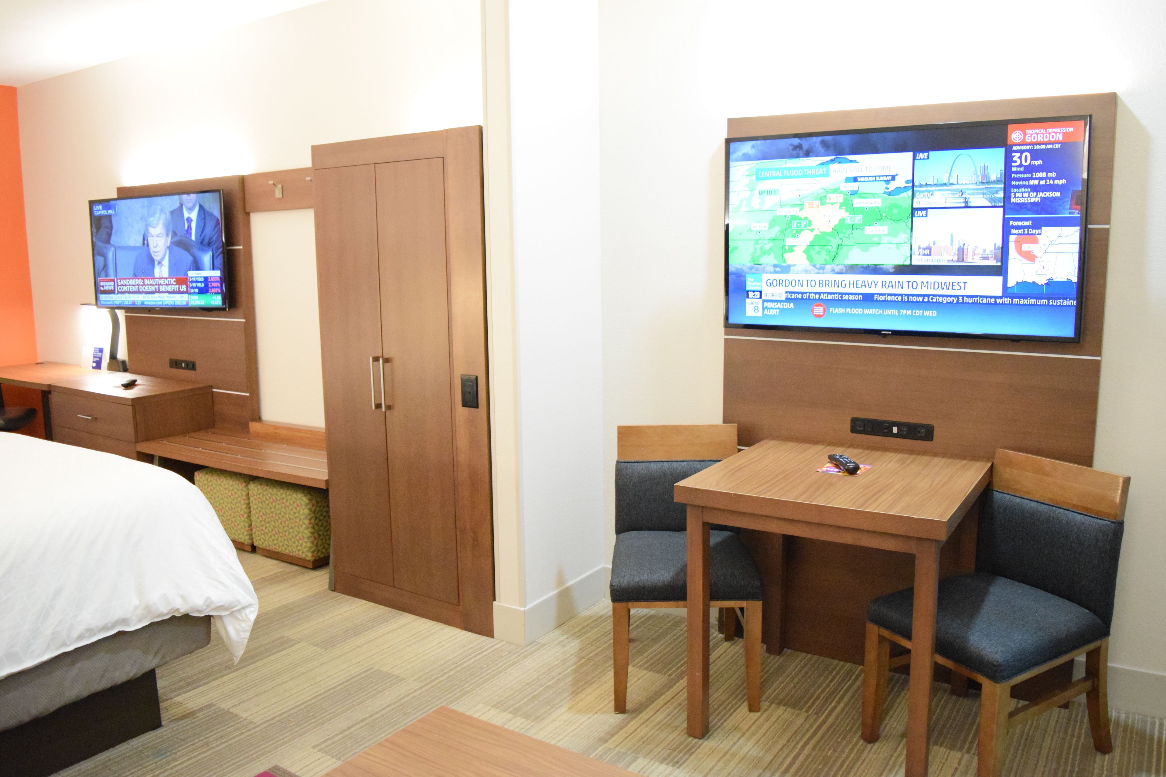 Habitación de hotel con cama, televisor montado en la pared, escritorio de madera con dos sillas y armario.