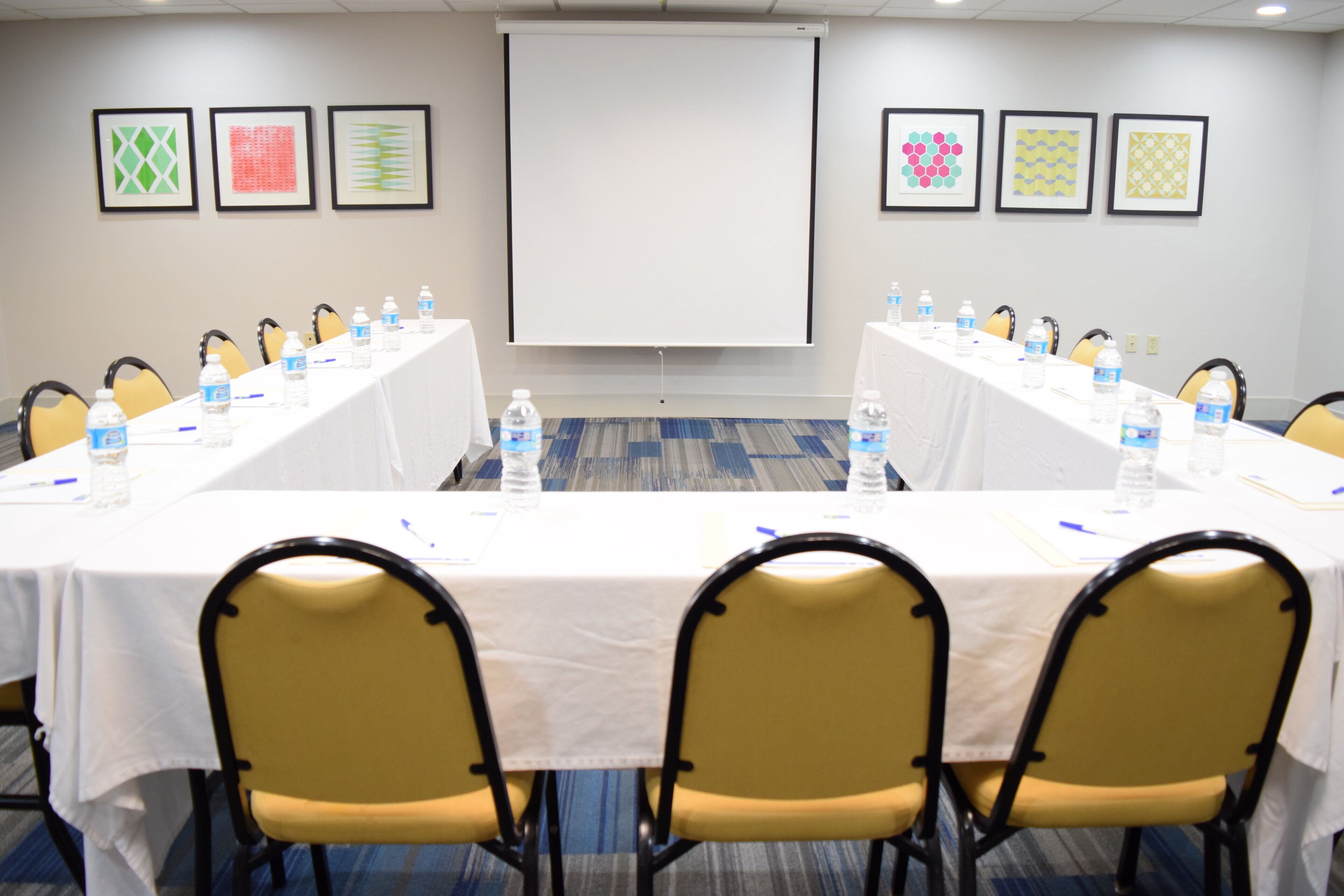 Sala de conferência com uma mesa comprida, cadeiras, garrafas de água e uma tela de projetor.