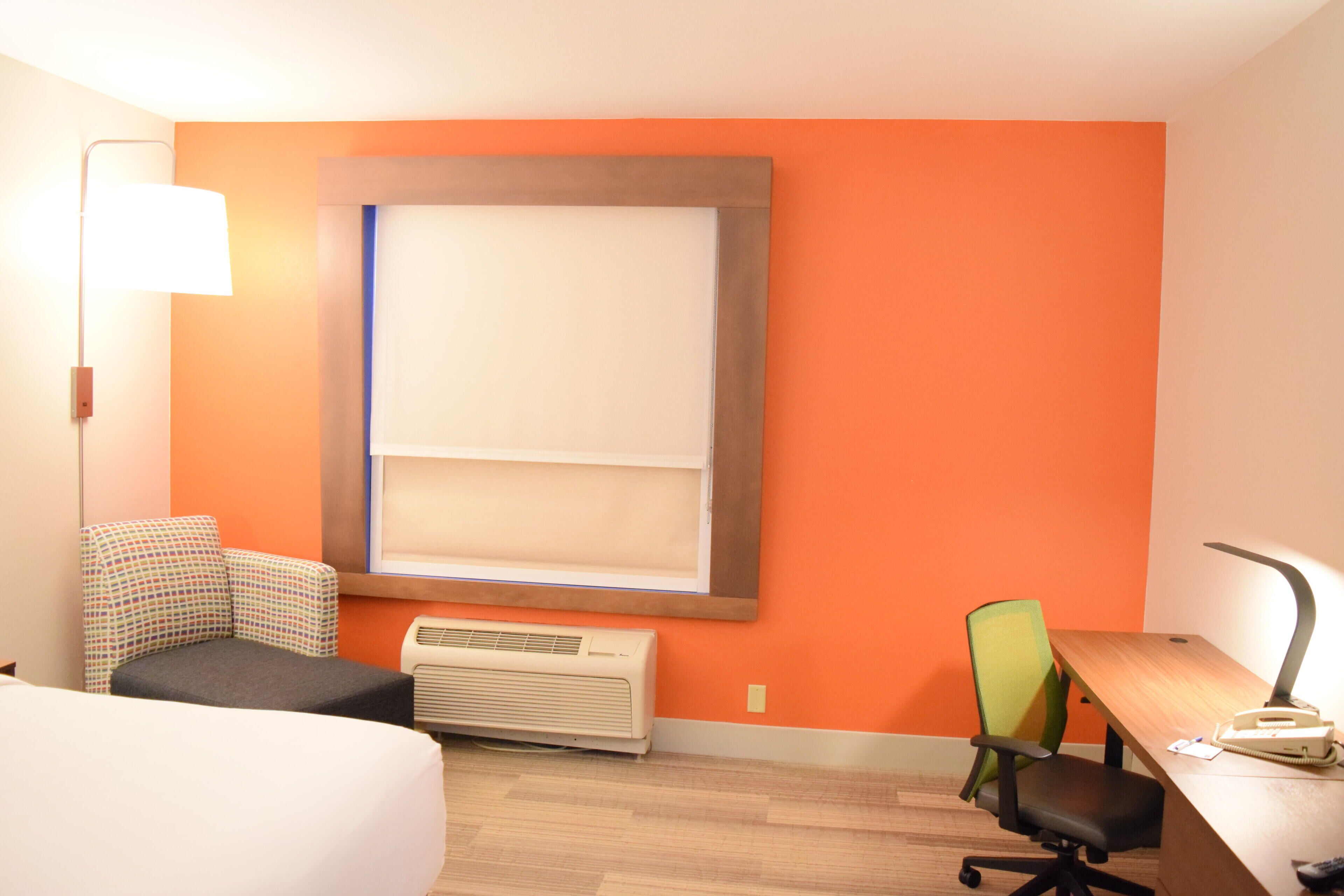 A hotel room with a bed, a desk with a chair, an air conditioner, a lamp, and a window with blinds.