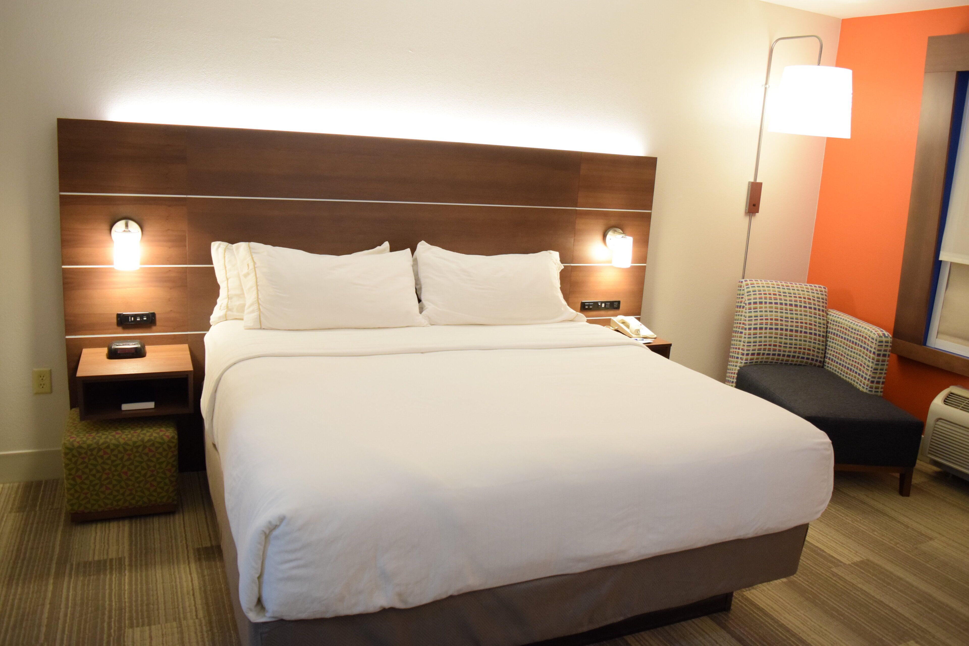 A hotel room with a large bed, bedside tables with lamps, a small chair, and a wall-mounted heater.