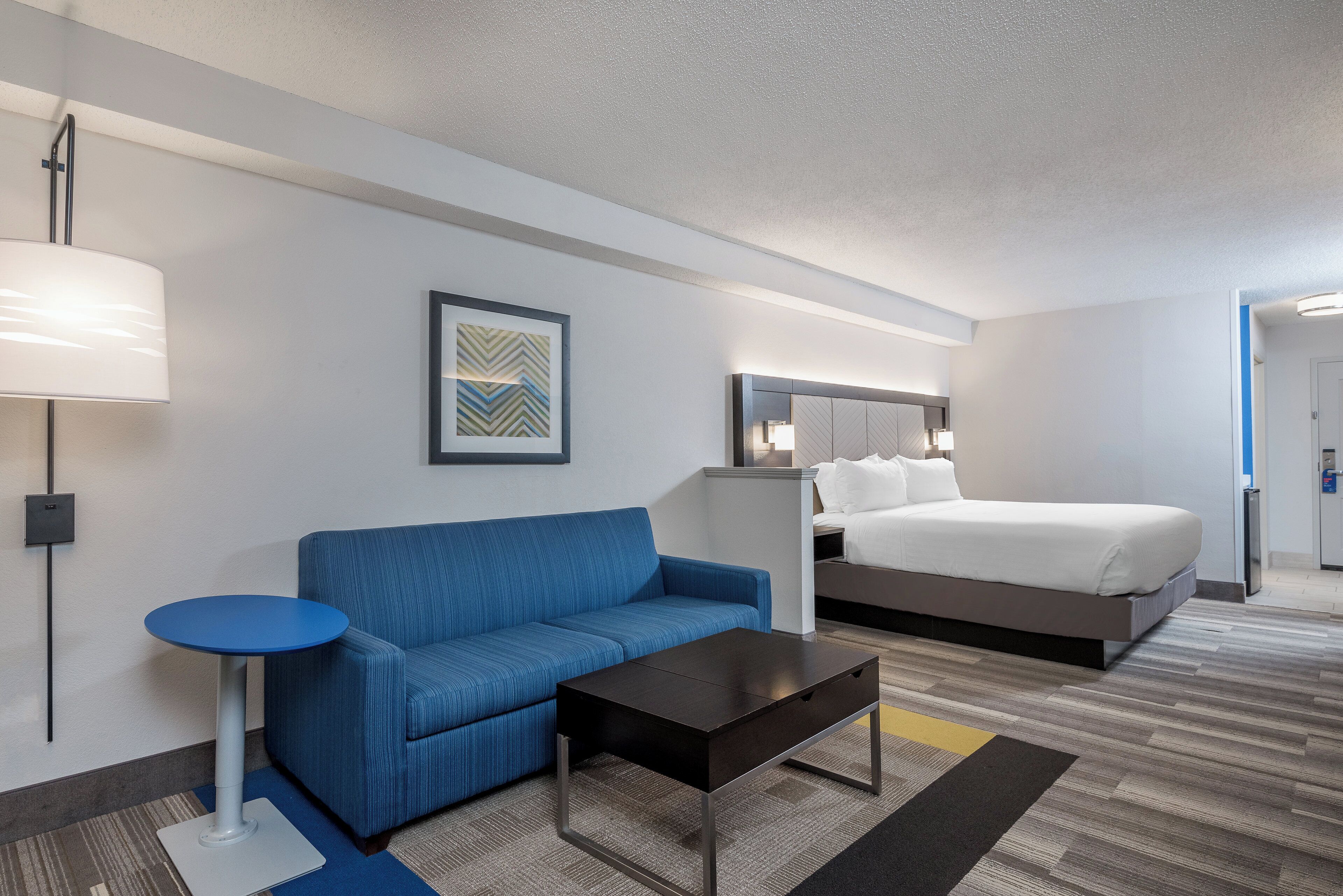 A hotel room with a bed, a blue sofa, a small table, a lamp, and a framed picture on the wall.