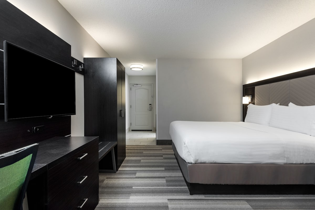 Una habitación de hotel moderna con un televisor de pantalla plana grande, una cama con ropa blanca, un aparador de madera oscura y piso alfombrado.