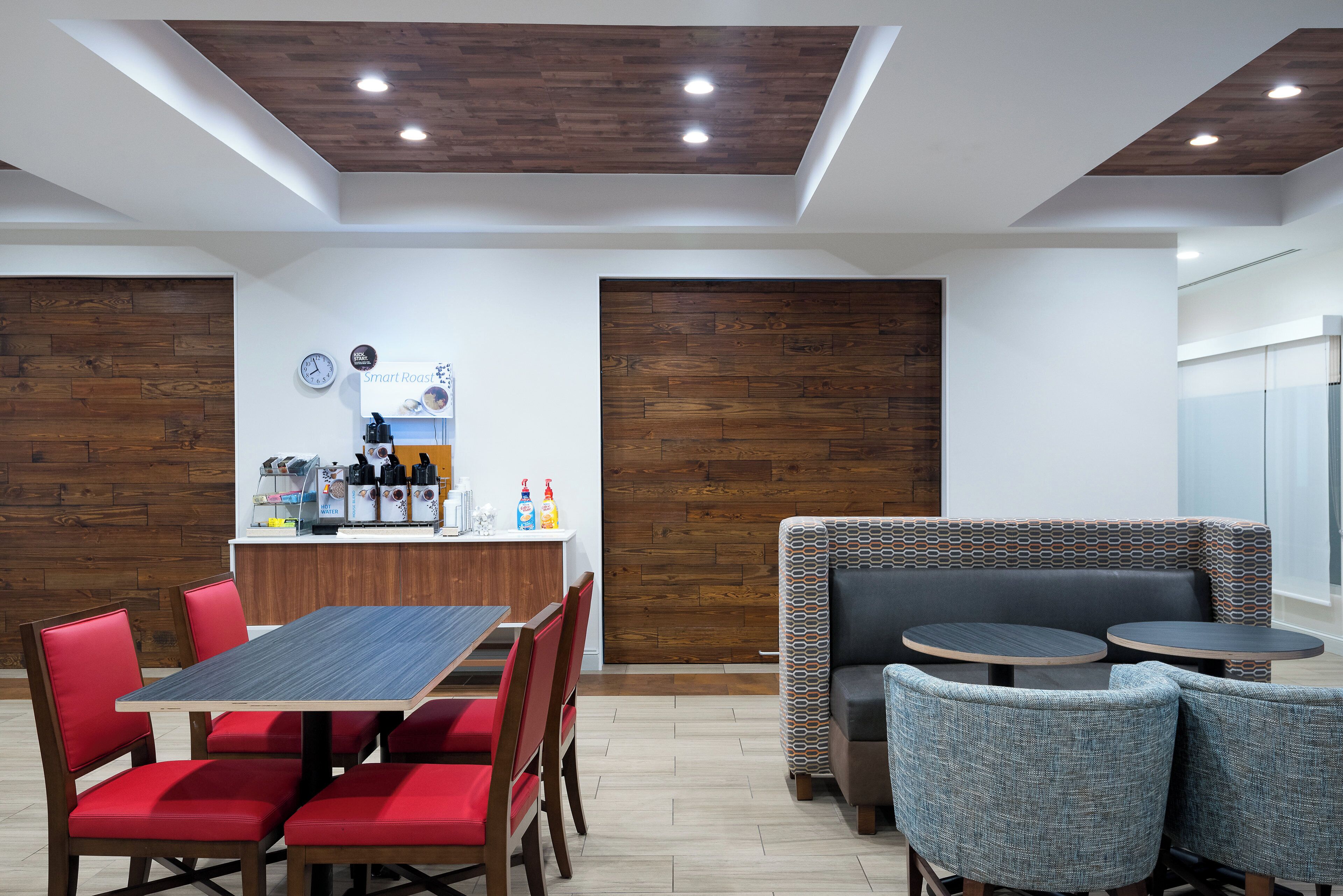 A modern cafe with a wooden ceiling, a central counter with a coffee machine, and seating arrangements including a booth and round tables.
