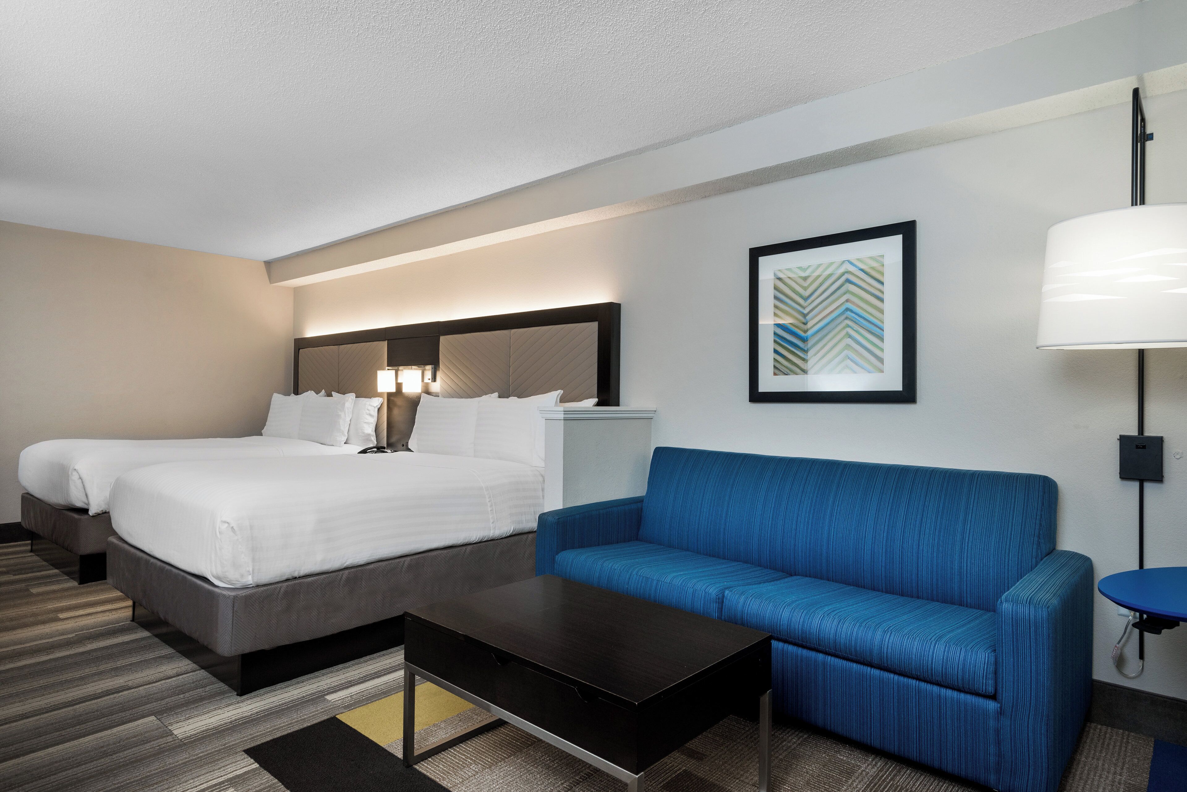 Habitación de hotel con dos camas, un sofá azul, una mesita y un cuadro en la pared.