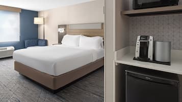 Camera d'albergo con un letto, un minibar, un forno a microonde e una zona salotto con una sedia e una lampada.