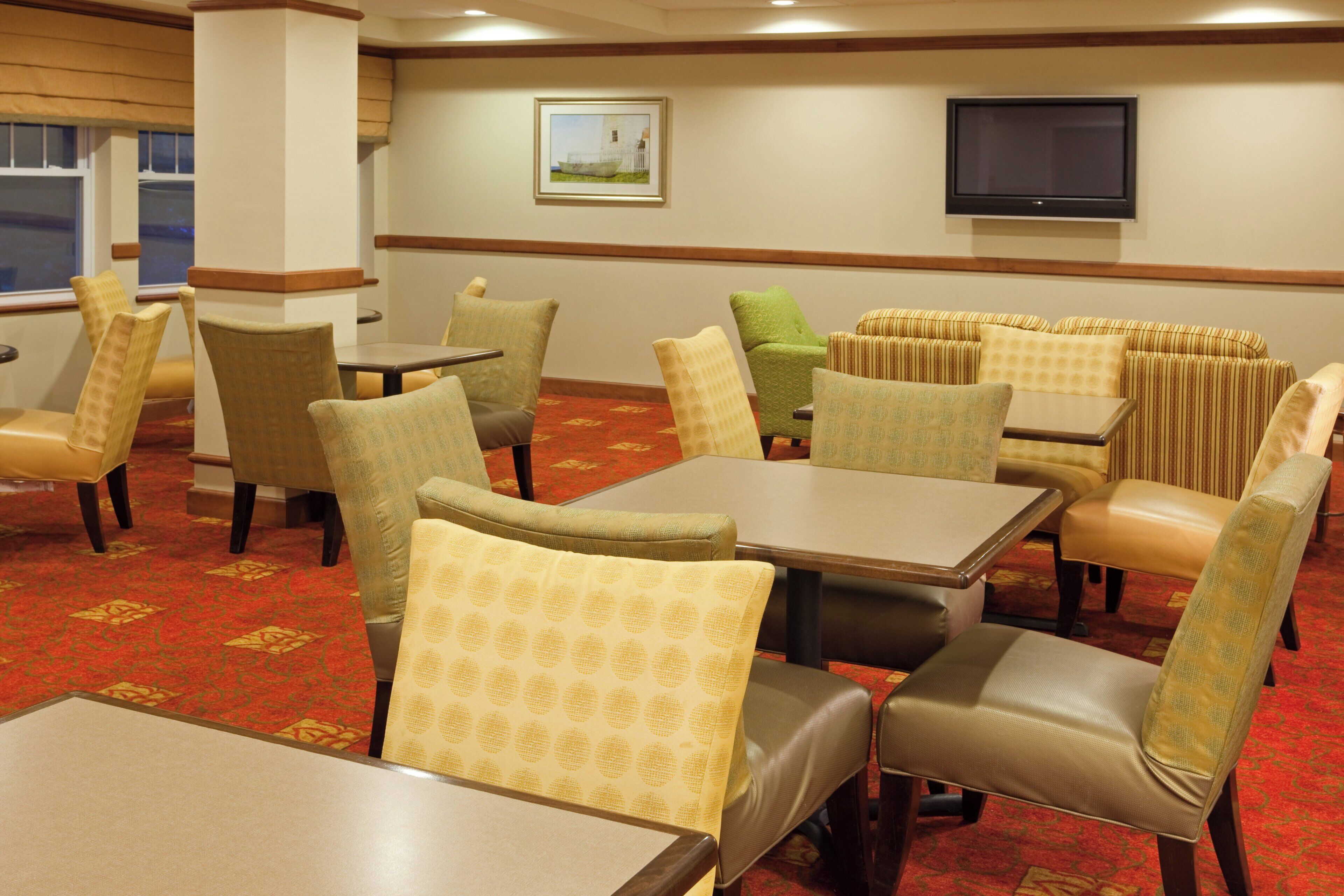 A hotel dining area with tables and chairs, a television on the wall, and a painting.