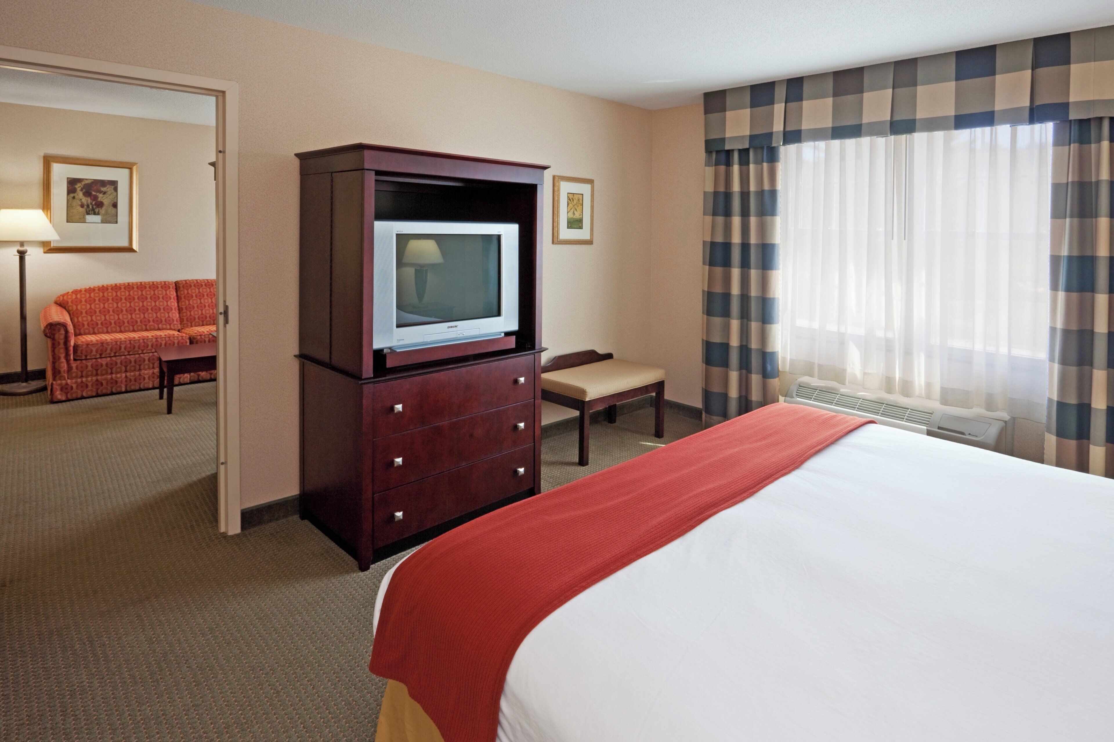 A hotel room with a bed, a television on a wooden cabinet, a chair, a desk, and a window with curtains.