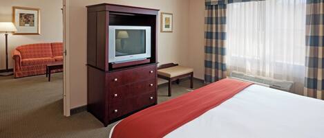 A hotel room with a bed, a television on a wooden cabinet, a chair, a desk, and a window with curtains.