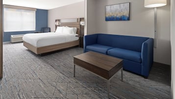 Habitación de hotel con cama, sofá azul, mesa de centro de madera, lámpara de pared y un cuadro.