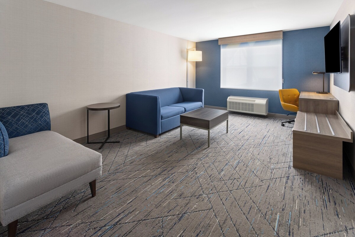 A hotel room with a blue sofa, a grey armchair, a small round table, a desk with a chair, a flat-screen TV mounted on the wall, and a large window with blinds.