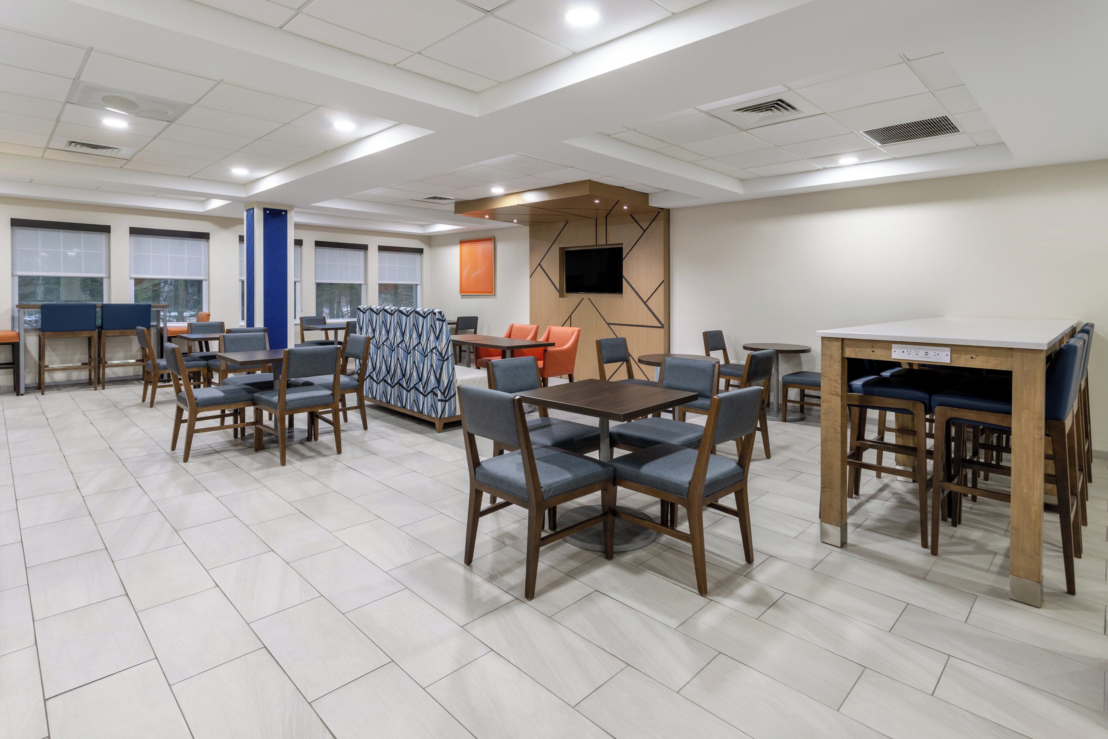 A spacious dining area with multiple tables and chairs, a television mounted on the wall, and large windows providing natural light.