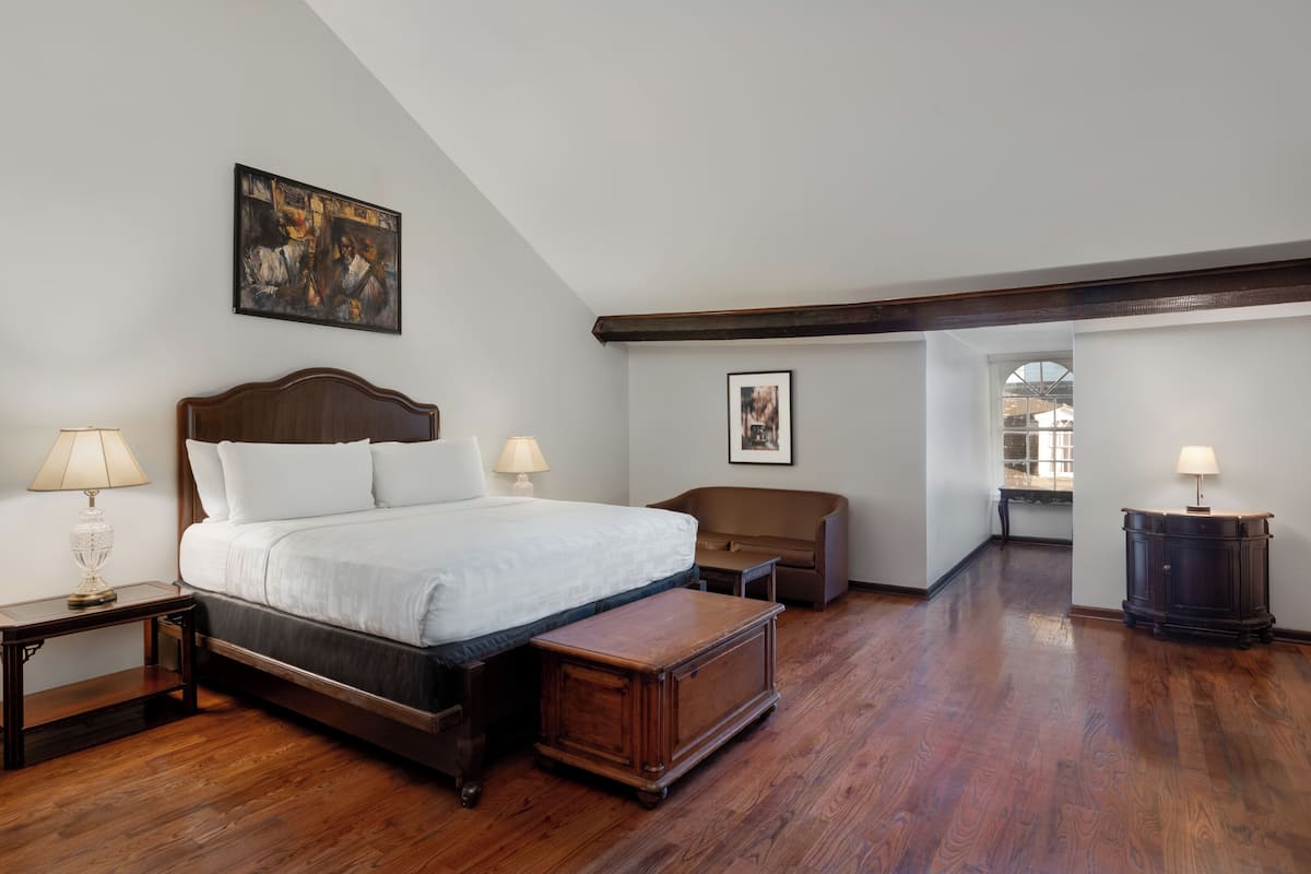 A bedroom with a large bed, a wooden nightstand, a bench, a dresser, and a painting on the wall.