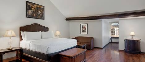 Un dormitorio con una cama grande, una mesita de noche de madera, un banco, un aparador y un cuadro en la pared.