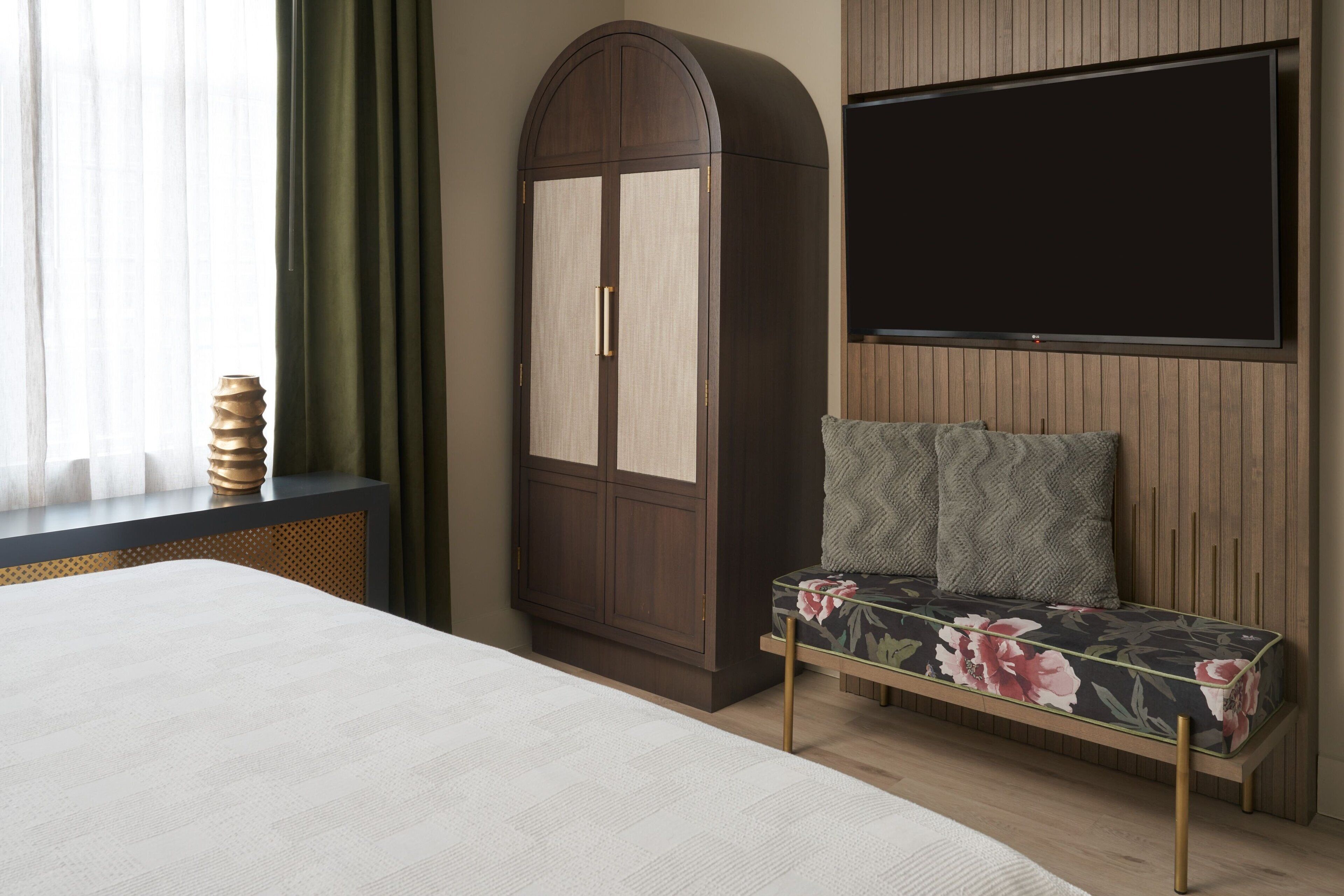A bedroom with a bed, a wardrobe, a bench with cushions, and a TV mounted on the wall.