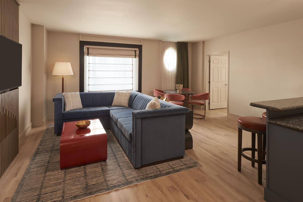 A modern living room with a sectional sofa, a red coffee table, a dining area with red chairs, and a kitchen counter.