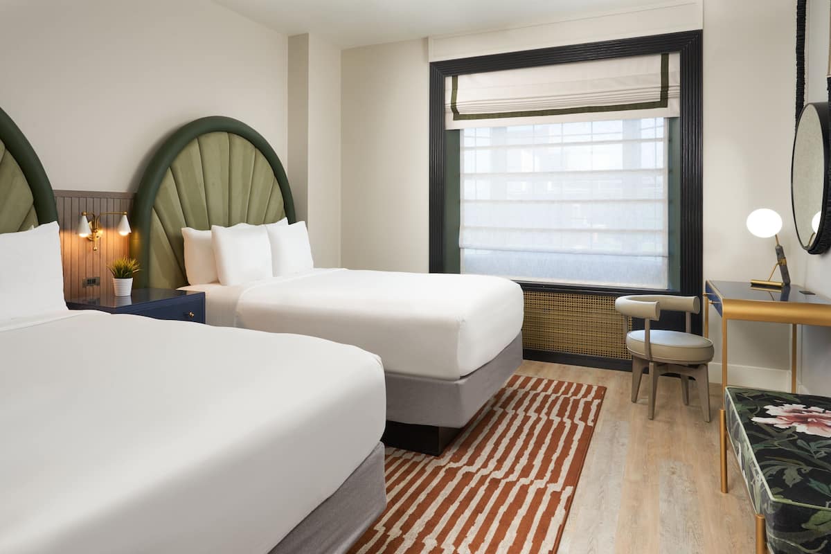 A hotel room with two beds, a desk, a chair, a large window with blinds, and a decorative mirror.