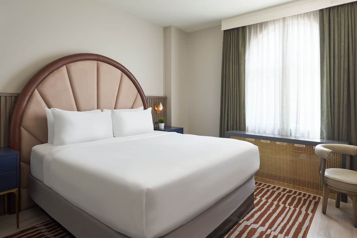 A large bed with a wooden headboard, a bedside table with a lamp, and a window with curtains.
