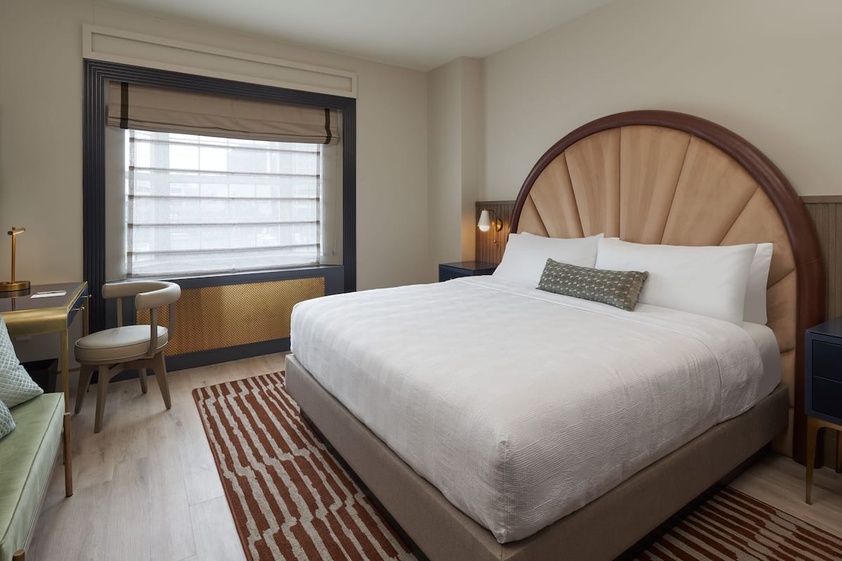 A hotel room with a large bed, a desk, a chair, and a window with blinds.