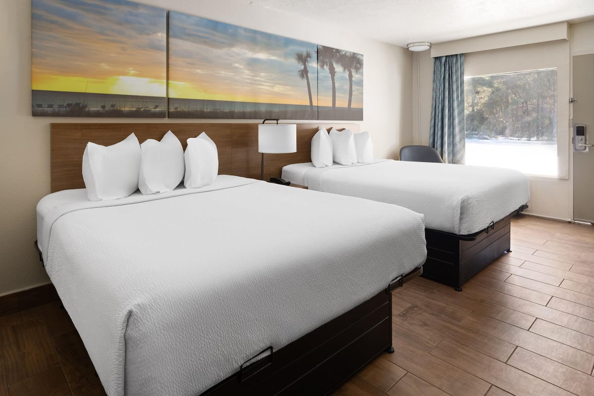 A hotel room with two beds, a nightstand, a desk, a chair, a large window with a view of trees, and a sunset painting on the wall.