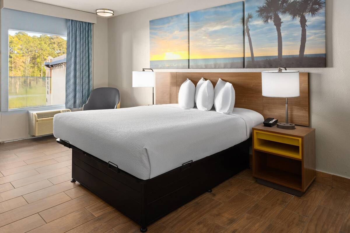 A hotel room with a large bed, a bedside table with a lamp, a chair, a window with a view of trees, and a painting of palm trees on the wall.