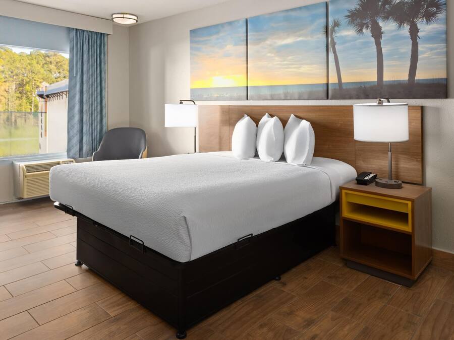 A hotel room with a large bed, a bedside table with a lamp, a chair, a window with a view of trees, and a painting of palm trees on the wall.