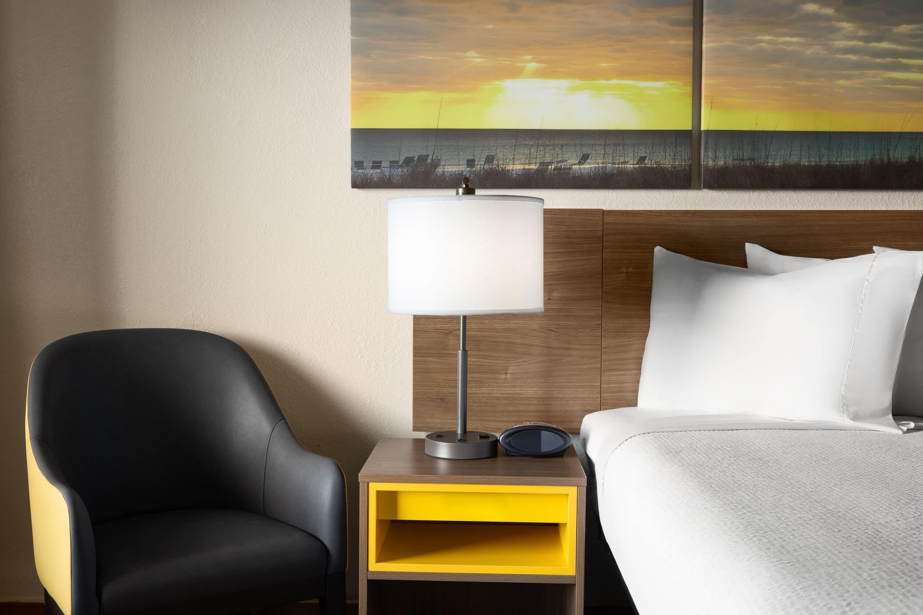 A hotel room with a bed, a nightstand with a lamp, a chair, and a painting of a sunset over the ocean.