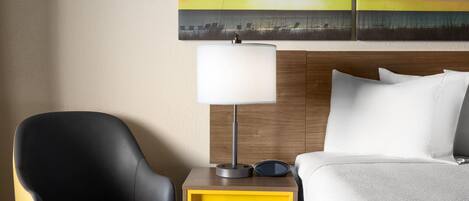 A hotel room with a bed, a nightstand with a lamp, a chair, and a painting of a sunset over the ocean.